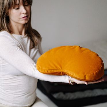 Kobieta prezentuje w dłoniach musztardową poduszkę do medytacji, jogi i relaksacji WellB w kształcie fasolki, na tle stosu złożonych mat.